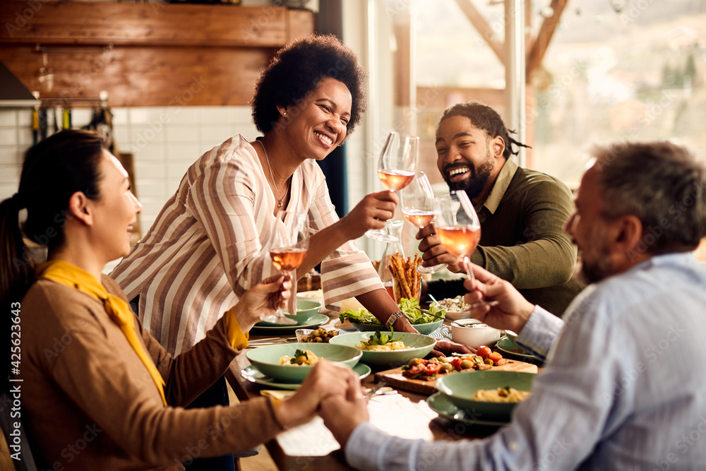 Multi-ethnic group of cheerful friends toasting with wine during meal in dining room.