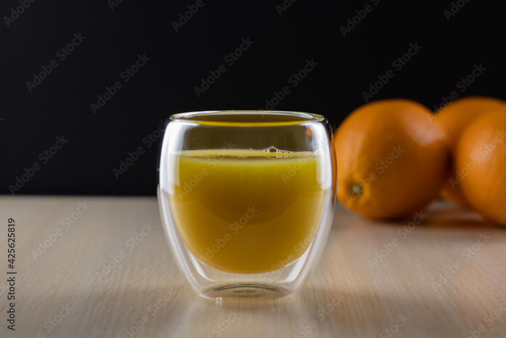 A glass with orange juice in the foreground on a black background. Whole oranges in the background. A glass with orange juice in the foreground on a black background. Whole oranges in the background.