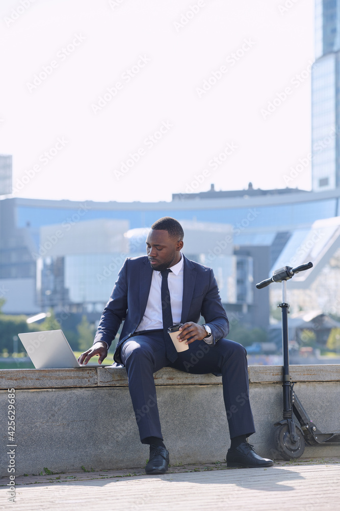 Young African businessman with laptop and coffee preparing presentation while sitting by riverside against skyscrapers and modern buildings Young African businessman with laptop and coffee preparing presentation while sitting by riverside against skyscrapers and modern buildings