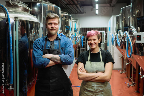 Two happy middle aged workers of beer production factory in aprons crossing arms by chest while standing in front of camera