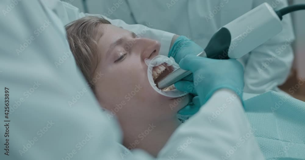 Close-up a male patient is lying in the dental chair and having his mouth open while male dentist is using intraoral scanner for examination oteeth and male dentist assistant is standing next to them. Close-up a male patient is lying in the dental chair and having his mouth open while male dentist is using intraoral scanner for examination oteeth and male dentist assistant is standing next to them.