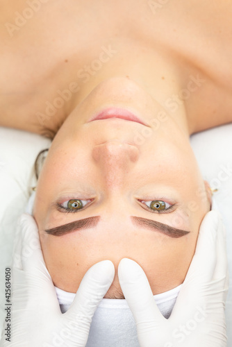 Specjalista sprawdza twarz młodej kobiety po zabiegu tatuowania brwi. Makijaż permanentny brwi w salonie urody. 