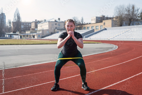  Young sportive woman trains legs with the help of fitness rubber bands on the red track. © Tania