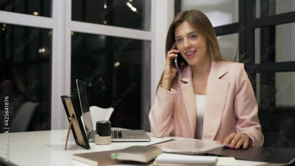 The young woman works till night. She speaks on the phone and looks at the documents. The young woman works till night. She speaks on the phone and looks at the documents.
