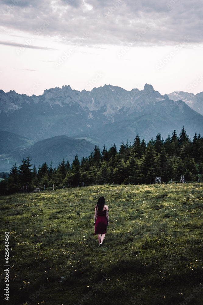 Back view of slim young woman walking on grassy valley on background of green mountains on cloudy evening