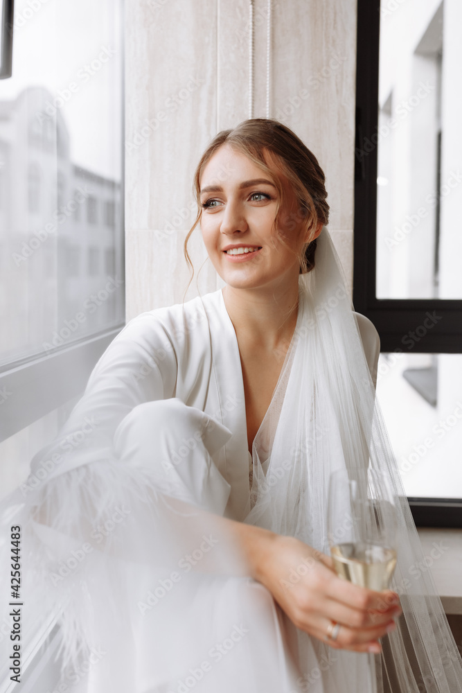Bride s morning. Bride drinking champagne in the peignoir. young woman is sitting on a large window in a hotel room in bathroom. Beautiful girl in white wedding robe. wedding day Bride s morning. Bride drinking champagne in the peignoir. young woman is sitting on a large window in a hotel room in bathroom. Beautiful girl in white wedding robe. wedding day