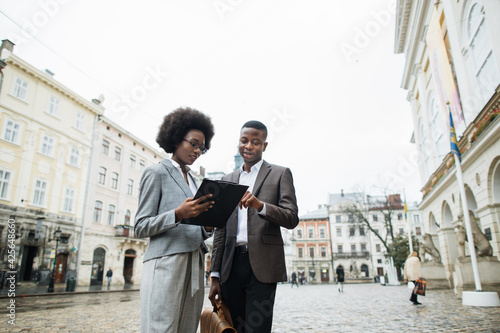 African male and female partners in business clothes standing on street and talking. Two colleagues communicating about work on fresh air.