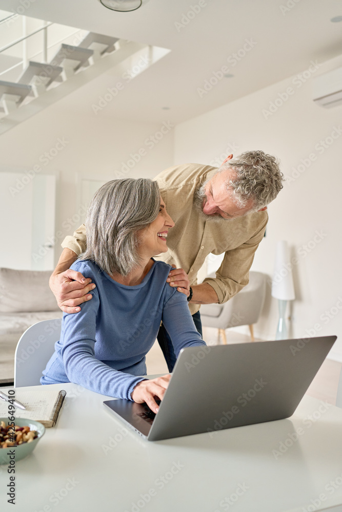 Obraz premium Happy mature senior 50s couple laughing using laptop computer at home. Smiling middle aged older husband embracing mid age wife having fun doing shopping online, working or browsing in living room.