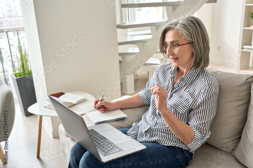 Mature adult old professional woman watching web training, online webinar on laptop computer remote working or e learning class from home office, video calling having virtual meeting, writing notes. photo