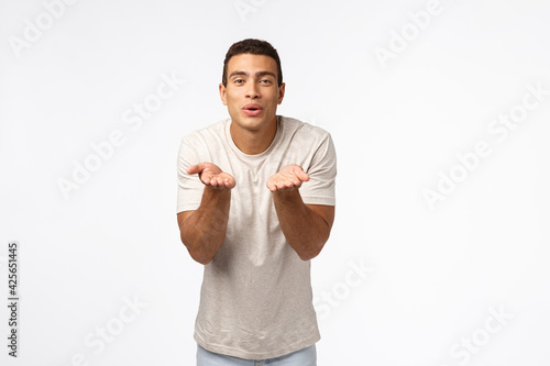 Cute, romantic pationate hispanic man in t-shirt, blowing kisses, fold lips smiling flirty and hold hands neart mouth, send mwah to camera, standing white background express affection or sympathy photo