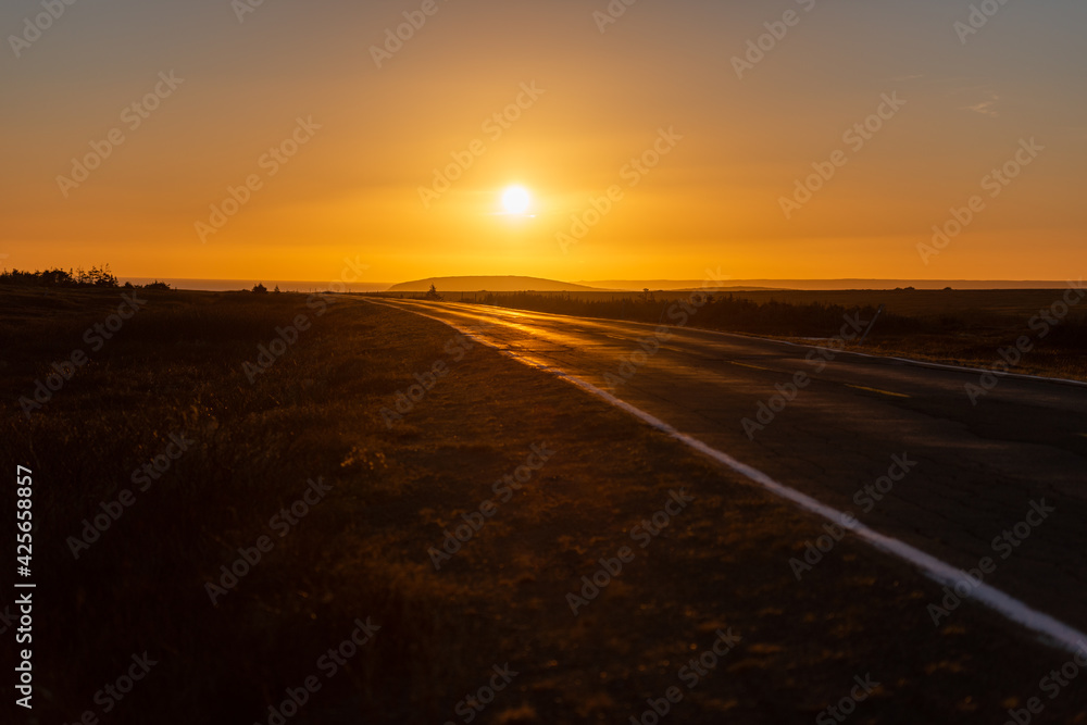 An inclined empty paved road with a white painted strip along the edge of the highway. The countryside is bare and barren. The sun is bright orange in the sky and is setting near the top of the road. 