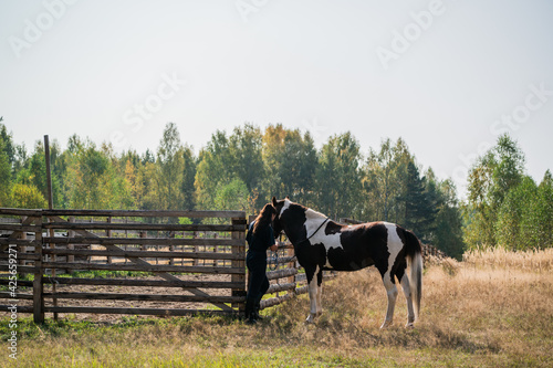 Young stable boy girl ties a brown horse with white spots to a fence
