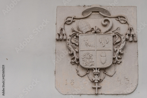 The old coat of arms of 1843 on the wall of the house. Coat of arms with papal hat, cross and plants.