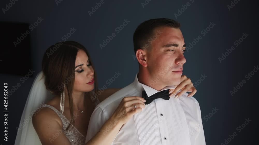 Bride helps groom get dressed and prepare for wedding ceremony. Couple is standing by the window and the wife puts a bow tie on her husband and he stands with his back to it for comfort. Portrait Bride helps groom get dressed and prepare for wedding ceremony. Couple is standing by the window and the wife puts a bow tie on her husband and he stands with his back to it for comfort. Portrait
