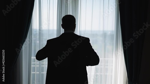 An elegant man stands by the window with curtains a day and puts on a jacket in preparation for an important event. Successful male businessman wearing a suit. Silhouette of a man behind