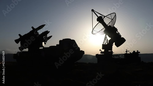 Creative artwork decoration. Silhouette of mobile air defence truck with radar antenna during sunset. Rocket launcher aimed at sky ready to attack. Selective focus