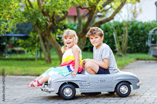 Two happy children playing with big old toy car in summer garden, outdoors. Kid boy pushing and driving car with little toddler girl, cute sister inside. Laughing and smiling kids. Lovely family © Irina Schmidt