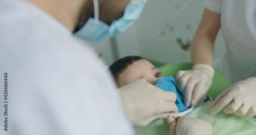 Close-up a little boy that laying in the dental chair with an open mouth and wearing a rubber dam on his teeth while the dentist is fixating the rubber dam by using white plastic detail. Close-up a little boy that laying in the dental chair with an open mouth and wearing a rubber dam on his teeth while the dentist is fixating the rubber dam by using white plastic detail.