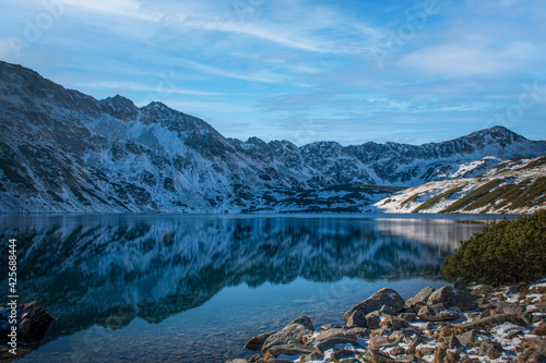 Symmetrical reflection on the surface of mountain lake water. Rocks on the shore, snow on the hills. December in Tatra Mountains. Selective focus on the ridge, blurred background. © juste.dcv