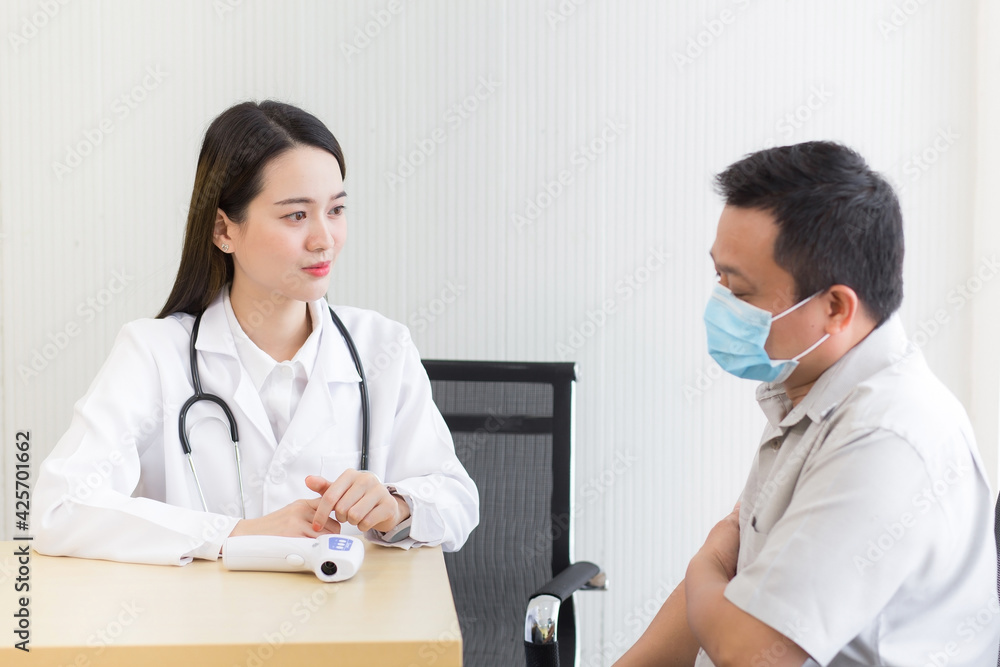 Asian beautiful woman doctor talking with a man patient about his pain and symptom while they put on a face mask to prevent Coronavirus disease and Thermometer on table in hospital.