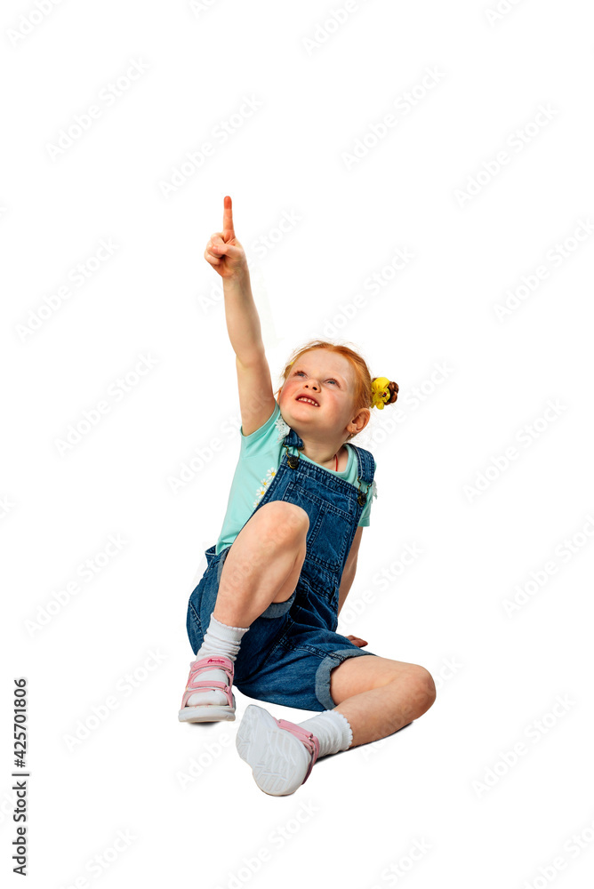 A little girl with red hair sits on a white background and shows her thumb up. Pointing gesture. A little girl with red hair sits on a white background and shows her thumb up. Pointing gesture.