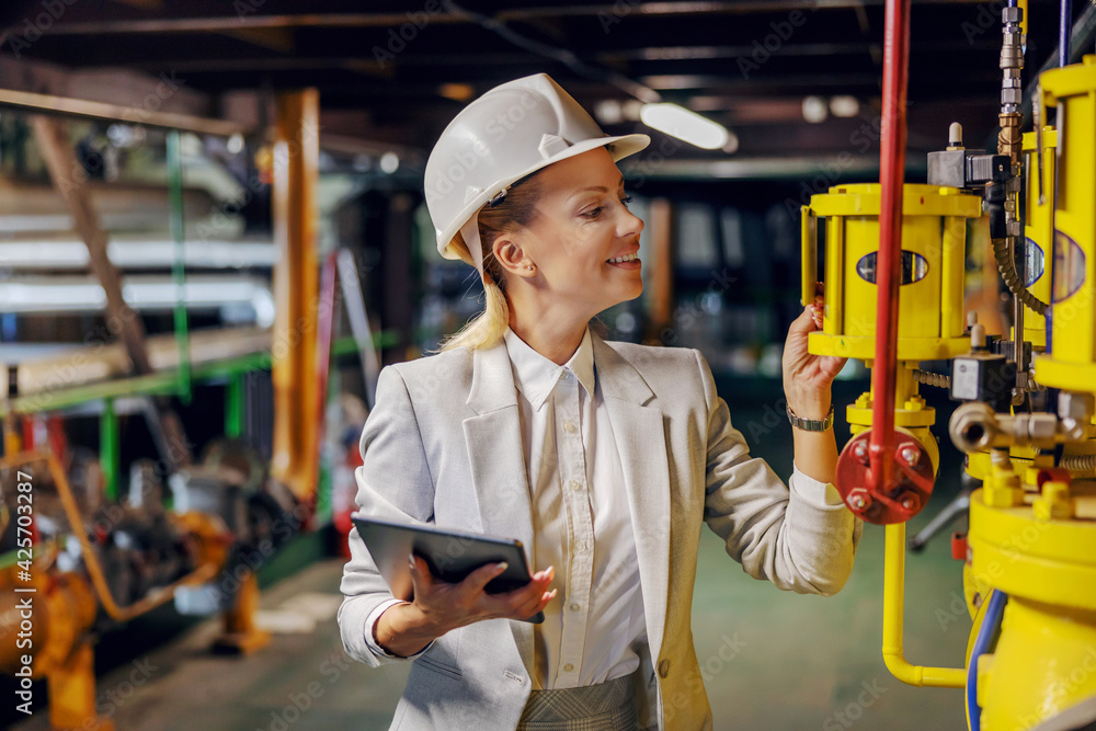 Blond dedicated successful businesswoman in formal wear checking on machinery and holding tablet while standing in power plant. Blond dedicated successful businesswoman in formal wear checking on machinery and holding tablet while standing in power plant.