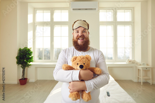 Happy cheerful adult grown up man with ginger beard and mustache in white pajamas and sleep mask smiling at camera standing in bedroom and cuddling teddy bear like a baby. Immature behavior concept photo
