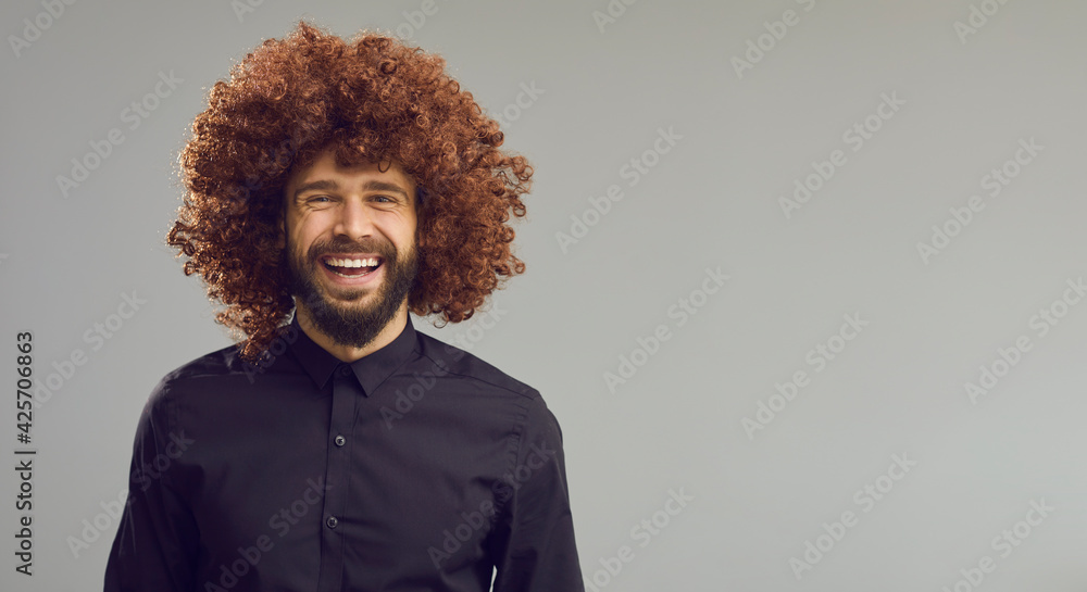 Obraz premium Happy young guy with thick curly brown hair on blank advertising space for text. Funny laughing man in Afro wig standing on light grey copy space banner background