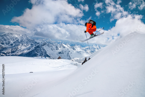A freerider skier in an orange suit with a backpack froze in a jump flight over high snow-capped peaks in the mountains on a sunny day