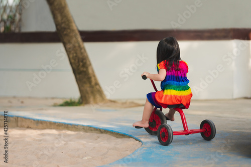Rear back view of cute girl riding red tricycle on playground blue road. Funny kid recreation. Active child wearing colorful dress. Along the road there is a sand pit. Happy children aged 4 years old.