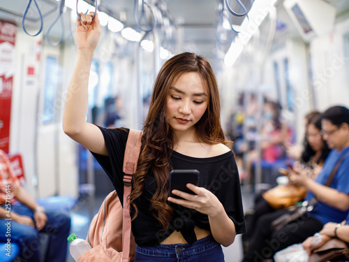 Картината върху платно Content Asian female riding train and chatting on social media via smartphone while holding handrail and looking at phone screen