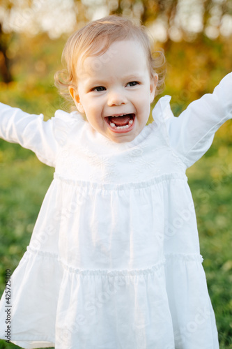 Joyful little girl child in a white cotton dress runs along a green meadow in the field.Healthy baby teeth in a 2-year-old child.