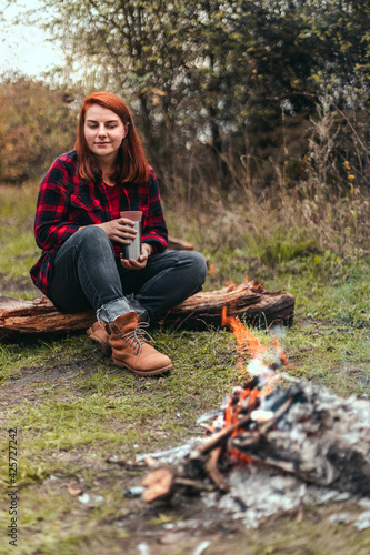 Female traveler camping in the woods and resting by the fire after a hard day. girl holding hot tea or coffee in the thermos. Trekking, adventure and seasonal vacation concept.