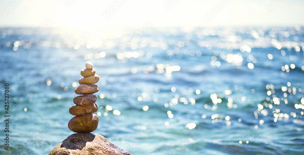 Obraz premium A pyramid of pebbles on a large stone against the background of the sea and the sun's glare on the water. Tour, tourism, vacation at the resort, swimming. Travel background, sunscreen. Copy space