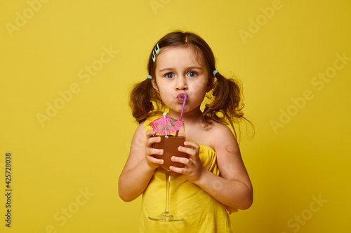 Wallpaper Mural Cute little girl drinking a tropical fruit cocktail from a straw, isolated on yellow background, copy space. Summer concept Torontodigital.ca