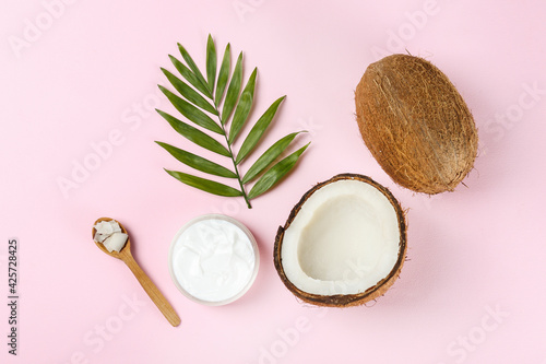 Coconut as a food source and cosmetic product. Whole and cracked fruit, a jar of moisturizing cream and oil, spoon and palm leaf on pink tabletop. Close up, top view, copy space, flat lay, background.
