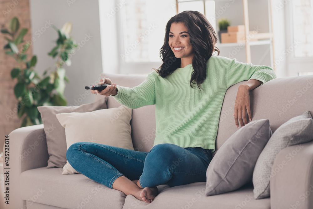 Full body photo of young afro woman happy positive smile sit sofa rest watch tv switch channel remote controller indoors Full body photo of young afro woman happy positive smile sit sofa rest watch tv switch channel remote controller indoors