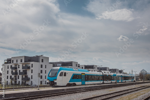 Modern white and blue passenger train on a station in front of a modern housing or residental district. City blocks and train.