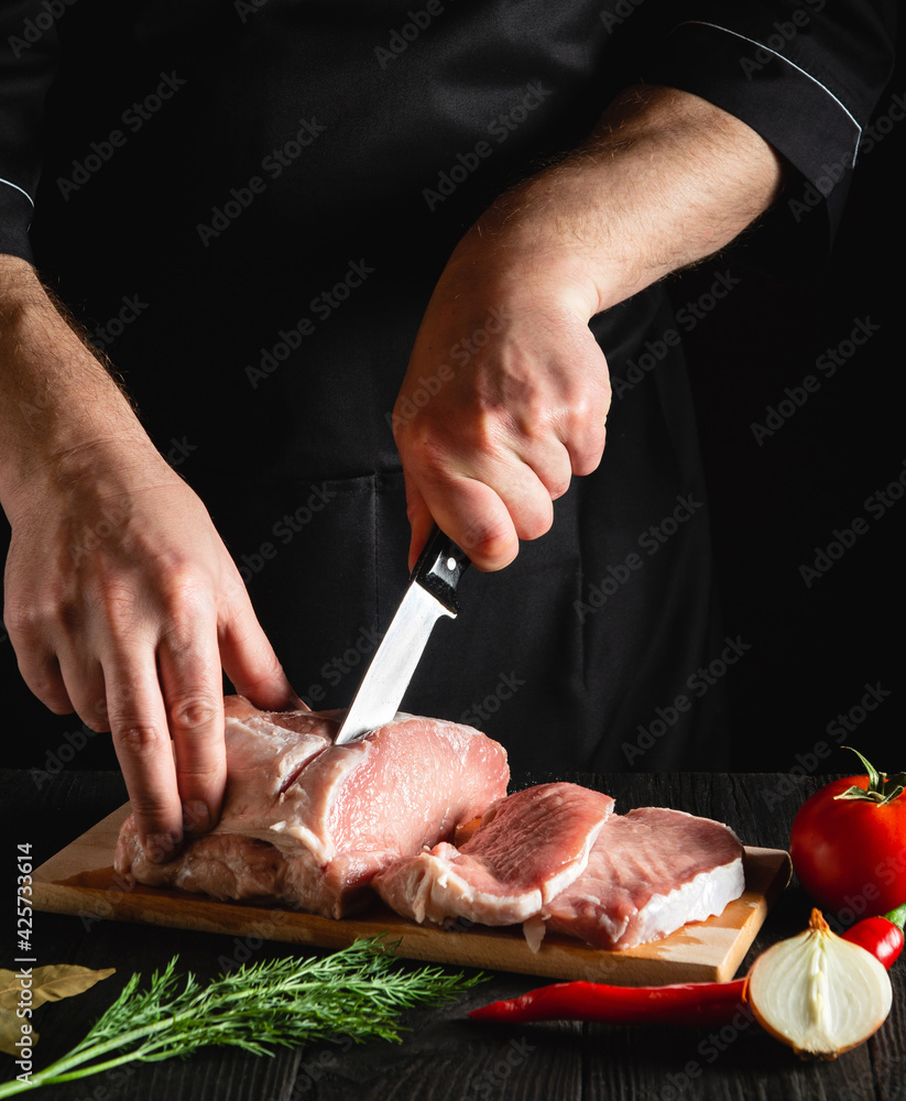 Chef cutting pork meat with knife on kitchen, cooking food. Vegetables and spices on the kitchen table to prepare delicious lunch