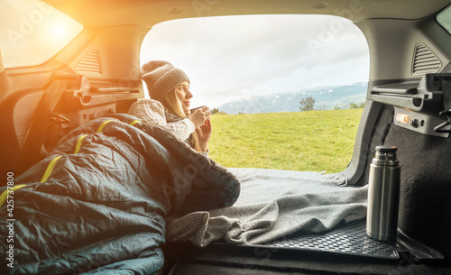 Girl resting in her car. Woman hiker, hiking backpacker traveler camper in sleeping bag, drinking hot tea and relaxing on top of mountain. Health care, authenticity, sense of balance and calmness. 