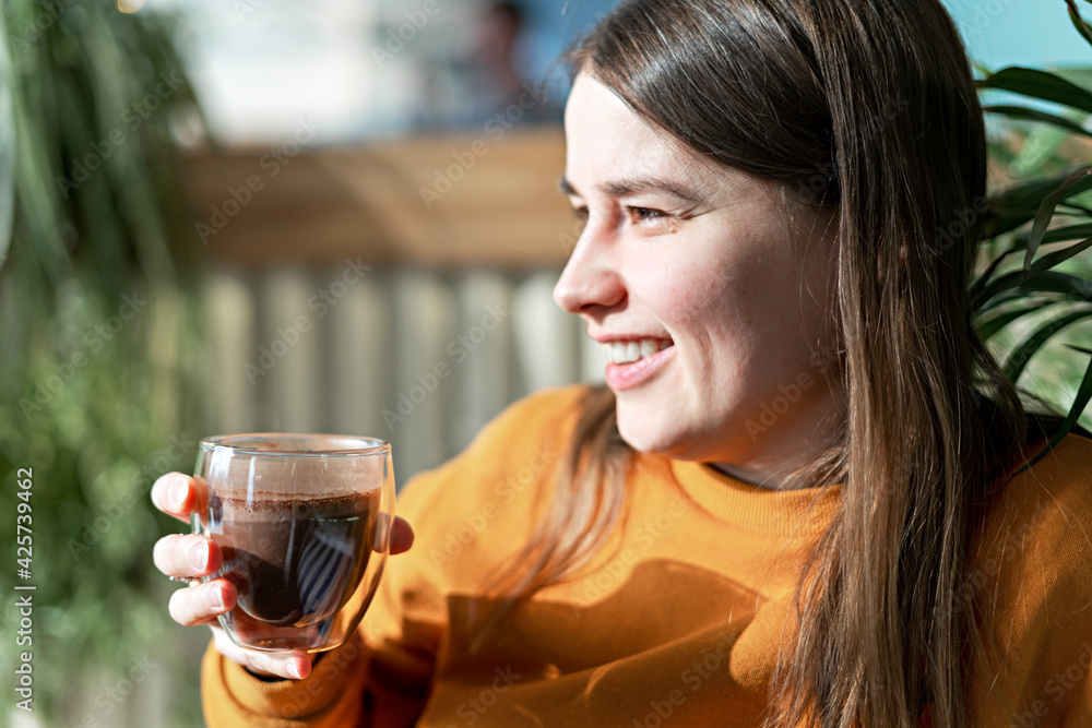 Smiling Young pretty Caucasian woman in orange sweatshirt holding glass cup of coffee or cocoa in sunlight on background of green house plants, coffeetime, hot drinks, hot chocolate Smiling Young pretty Caucasian woman in orange sweatshirt holding glass cup of coffee or cocoa in sunlight on background of green house plants, coffeetime, hot drinks, hot chocolate