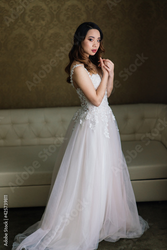 Asian bride in wedding dress in hotel sitting on the bed, standing near window, morning