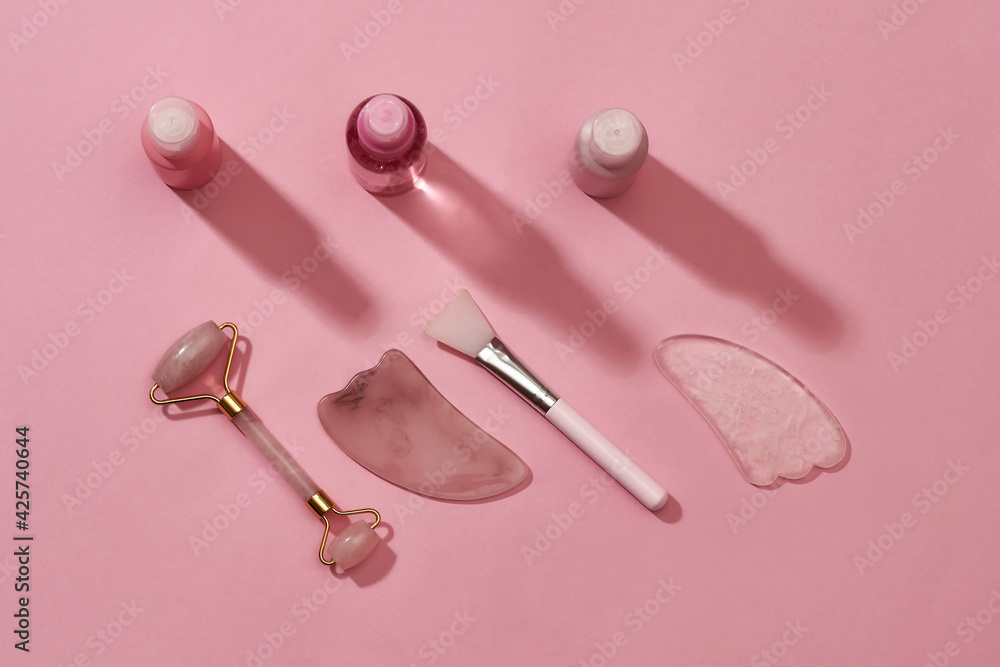 Flat lay of rose quartz facial massage tools such as gua sha stone scrapers, face roller and beauty products in plastic bottles isolated on pink surface Flat lay of rose quartz facial massage tools such as gua sha stone scrapers, face roller and beauty products in plastic bottles isolated on pink surface