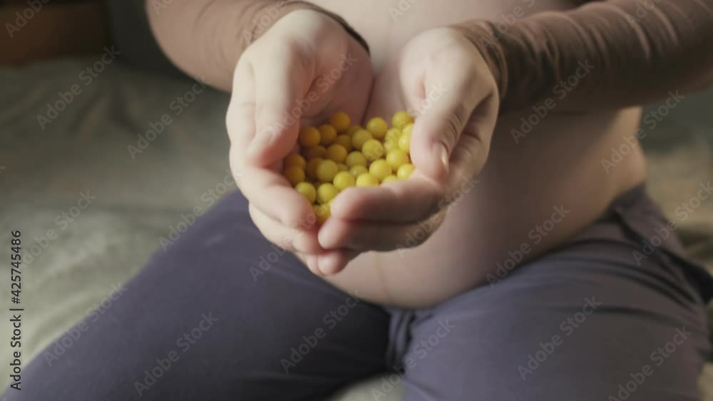pregnant woman sitting on couch, holding bunch of yellow vitamins in palms pregnant woman sitting on couch, holding bunch of yellow vitamins in palms