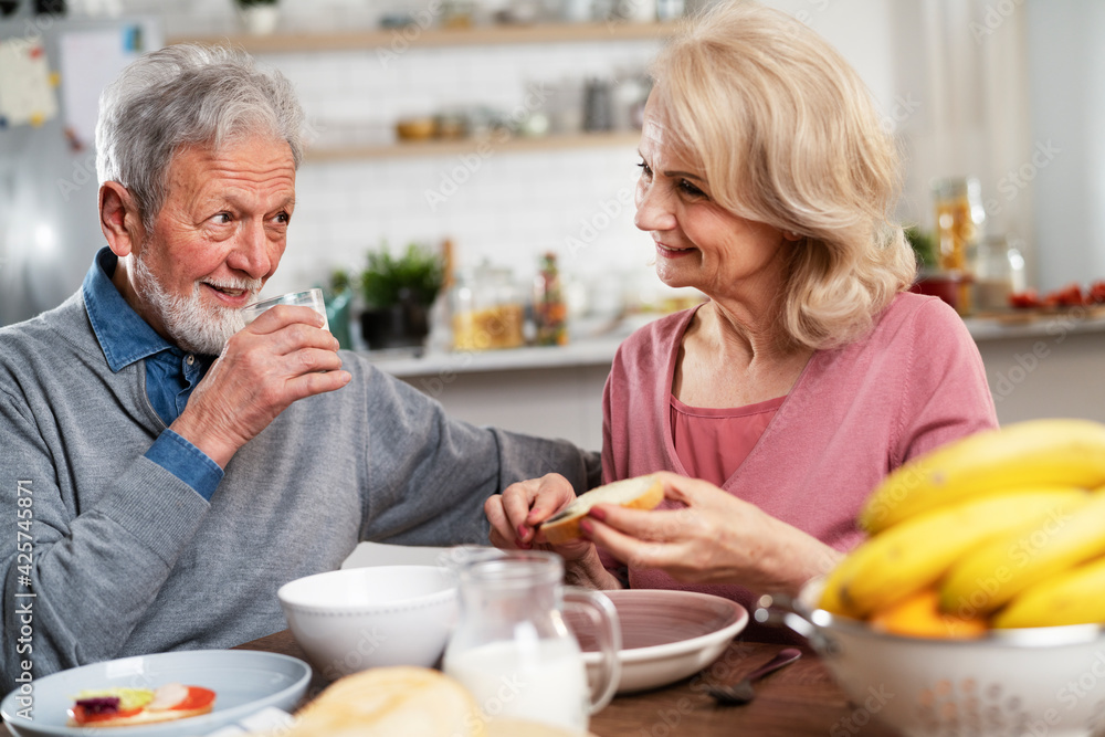 Senior couple eating breakfast in the kitchen. Husband and wife talking and laughing while eating a sandwich Senior couple eating breakfast in the kitchen. Husband and wife talking and laughing while eating a sandwich