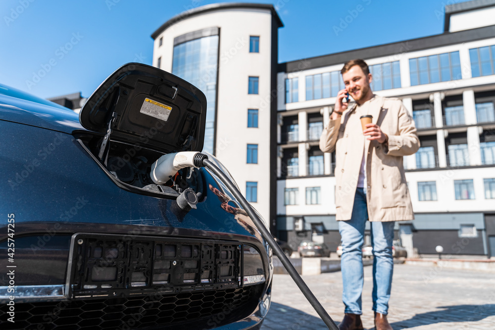 Obraz premium Stylish man talks on the phone and drinks coffee while his car is charging at the station