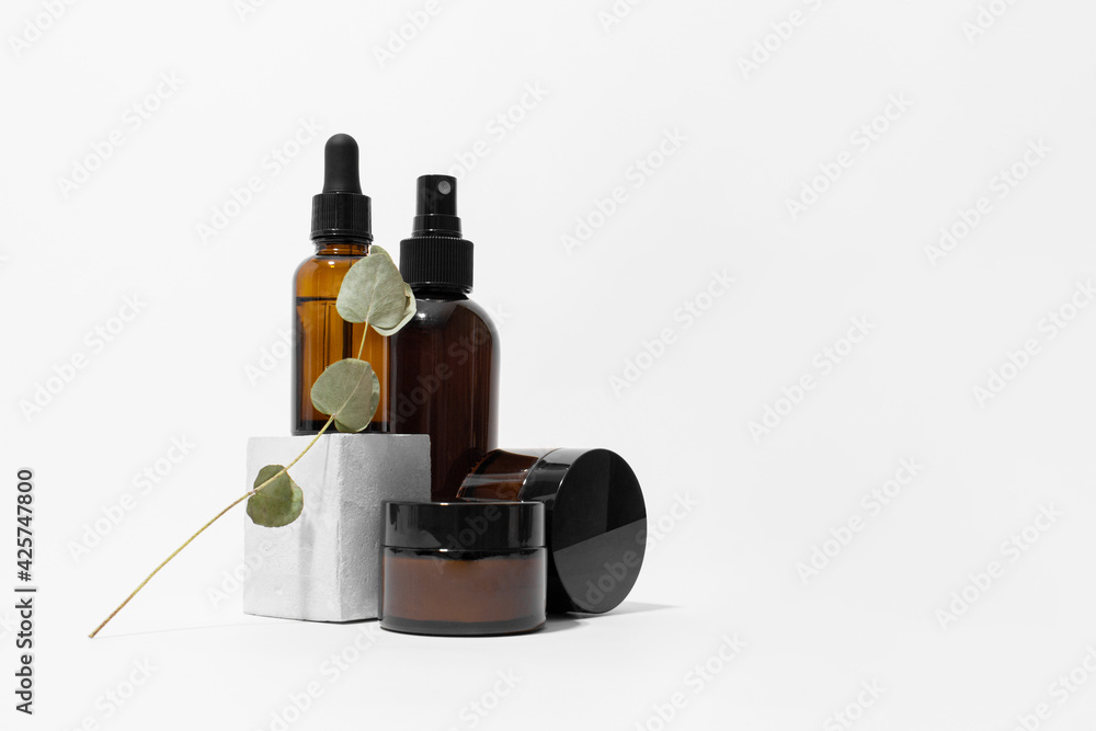 Brown dropper glass pipette and spray bottles and jars on concrete pedestal with eucalyptus, white background, copy space. Cream, serum, oil on podium. Natural cosmetic. Beauty, skincare product
