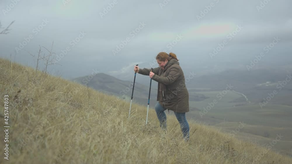Fat middle aged woman climbing a hill using Nordic walking sticks against the backdrop of an mountain landscape Fat middle aged woman climbing a hill using Nordic walking sticks against the backdrop of an mountain landscape