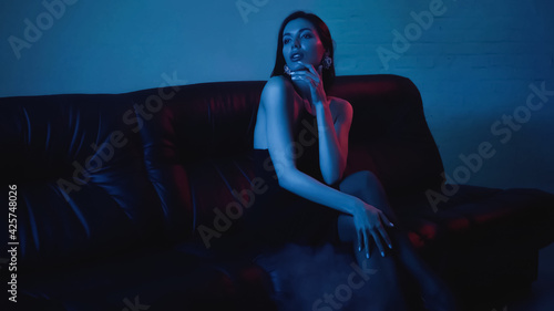 red lighting on face of pensive brunette woman sitting on black couch on blue
