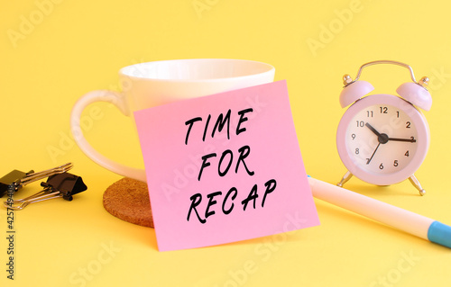 Pink paper with the text TIME FOR RECAP on a white cup. Clock, pen on a yellow background.